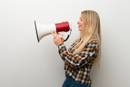 Blonde young girl over white wall shouting through a megaphone to announce something in lateral positionの写真素材