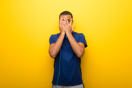 African american man with blue t-shirt on yellow background covering eyes by hands and looking through the fingersの写真素材