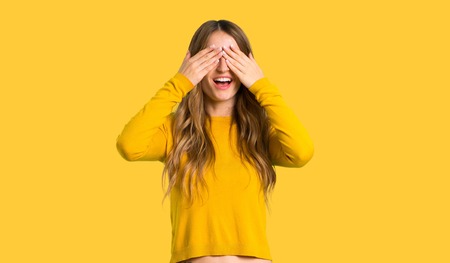 young girl with yellow sweater covering eyes by hands. Surprised to see what is ahead on isolated yellow backgroundの写真素材