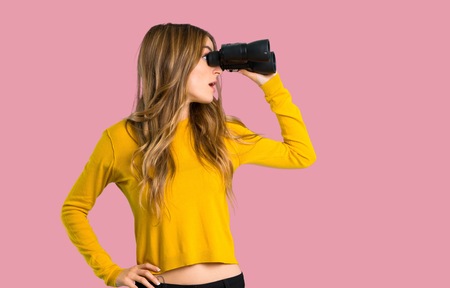 young girl with yellow sweater and looking in the distance with binoculars on isolated pink backgroundの写真素材