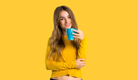 young girl with yellow sweater holding a hot cup of coffee on isolated yellow backgroundの写真素材