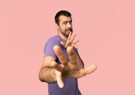 Handsome man is a little bit nervous and scared stretching hands to the front on isolated pink...