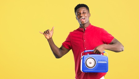 Young african american man holding a blue vintage radio while singing on yellow backgroundの写真素材