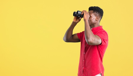 Young african american man and looking for something in the distance with binoculars on yellow...