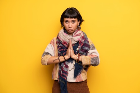 Young hippie woman over yellow wall keeps palm together. Person asks for somethingの写真素材