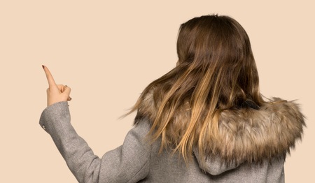 Teenager girl with coat pointing back with the index finger on isolated yellow backgroundの写真素材