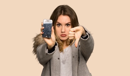 Teenager girl with coat with a broken phone on isolated yellow backgroundの写真素材