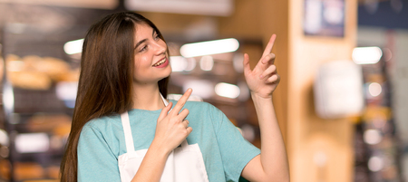 Girl with apron pointing with the index finger and looking up in a bakeryの写真素材