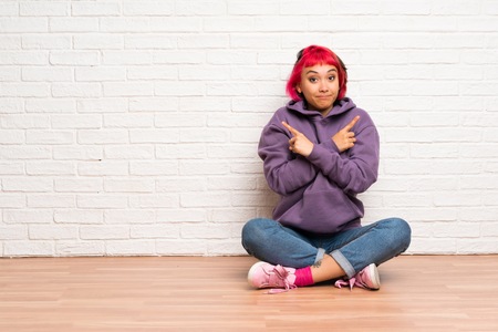 Young woman with pink hair sitting on the floor pointing to the laterals having doubtsの写真素材