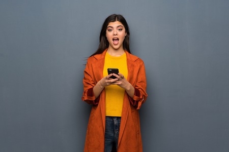Teenager girl with coat over grey wall surprised and sending a messageの写真素材