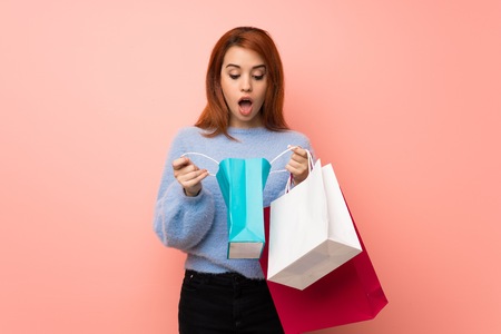 Young redhead woman over pink background surprised while holding a lot of shopping bagsの写真素材