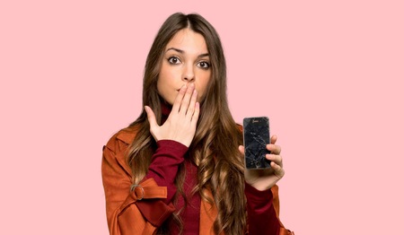 Young woman with coat with troubled holding broken smartphone over isolated pink background