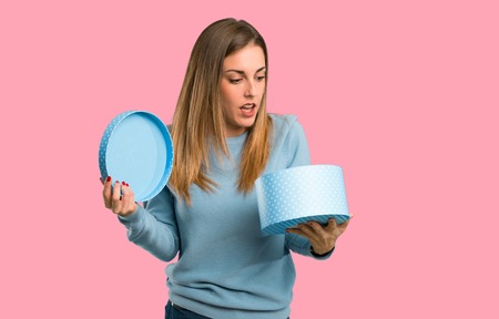Blonde woman with blue shirt holding gift box in hands on isolated pink backgroundの写真素材