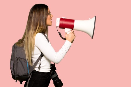 Young photographer woman shouting through a megaphone on isolated pink background