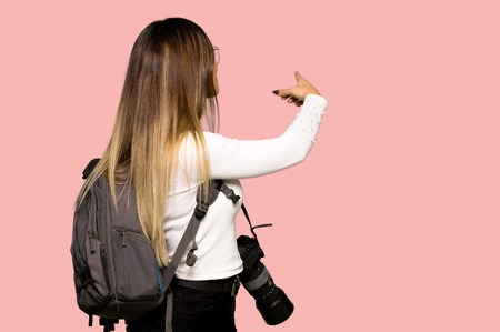 Young photographer woman pointing back with the index finger on isolated pink backgroundの写真素材