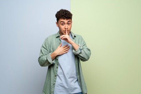 Young man over blue and green background showing a sign of silence gesture putting finger in mouthの写真素材