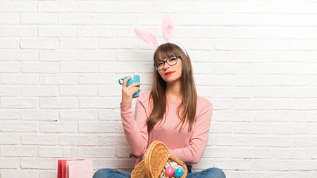 Woman with bunny ears for Easter holidays sitting on the floor taking a coffee in takeaway paper cup...