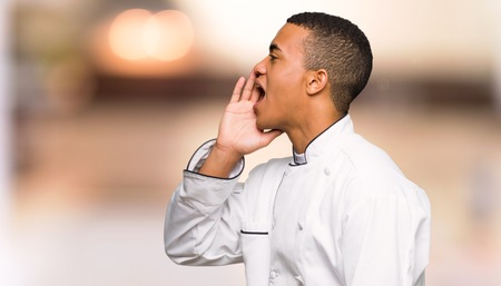 Young afro american chef man shouting with mouth wide open to the lateral on unfocused backgroundの写真素材