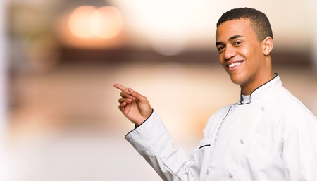 Young afro american chef man pointing finger to the side in lateral position on unfocused backgroundの写真素材