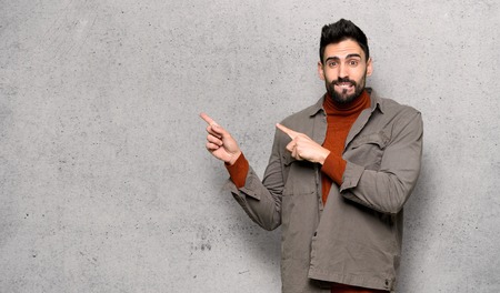 Handsome man with beard frightened and pointing to the side over textured wallの写真素材
