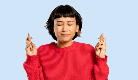 Short hair girl with red sweater with fingers crossing and wishing the best on isolated blue...
