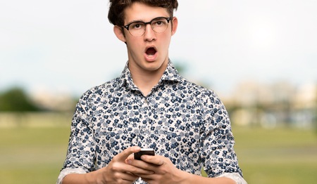 Teenager man with flower shirt and glasses surprised and sending a message at outdoorsの写真素材
