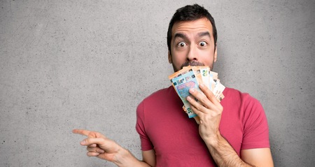 Man taking a lot of money pointing finger to the side with a surprised face over textured wall