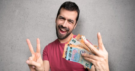 Man taking a lot of money smiling and showing victory sign with both hands over textured wallの写真素材