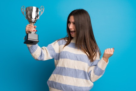 Young woman over blue wall holding a trophyの写真素材
