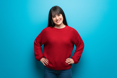 Woman with red sweater over blue wall posing with arms at hip and smilingの写真素材