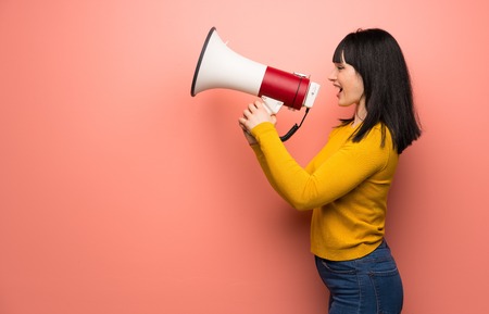 Woman with yellow sweater over pink wall shouting through a megaphone