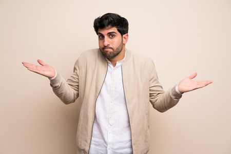 Young man over isolated wall having doubts while raising hands