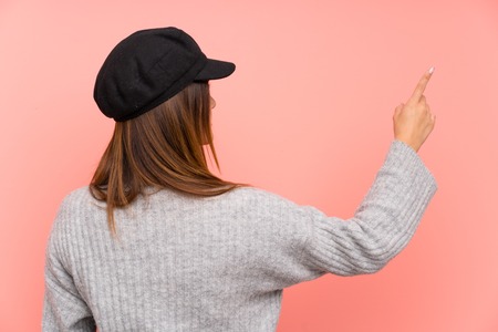Fashion woman with hat over pink wall pointing back with the index fingerの写真素材