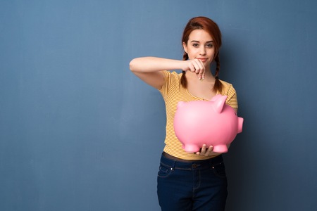 Young redhead woman over blue background taking a piggy bank and happy because it is fullの写真素材
