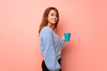 Young redhead woman over pink background holding a hot cup of coffeeの写真素材