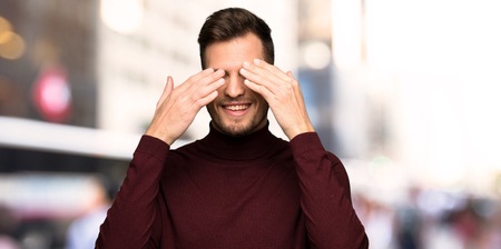 Man with turtleneck sweater covering eyes by hands. Surprised to see what is ahead in the cityの写真素材