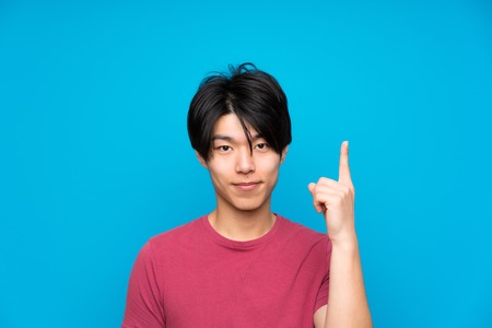 Asian man with red shirt over isolated blue wall pointing with the index finger a great ideaの写真素材