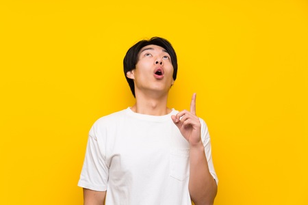 Asian man over isolated yellow wall pointing up and surprisedの写真素材