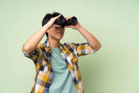 Asian man over isolated green wall with black binocularsの写真素材
