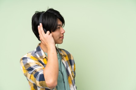 Asian man over isolated green wall listening to music with headphonesの写真素材