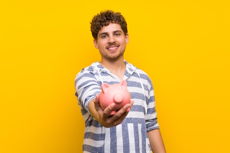 Blonde man over yellow wall holding a piggybankの写真素材