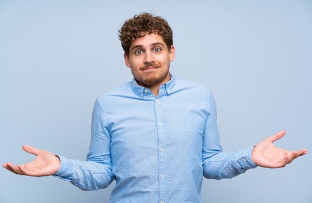 Blonde man over blue wall having doubts while raising handsの写真素材