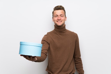Blonde man over isolated white wall holding a gift in hands