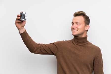Blonde man over isolated white wall holding a camera