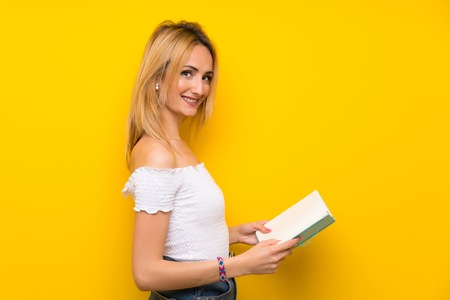 Young blonde woman over isolated yellow wall holding and reading a book