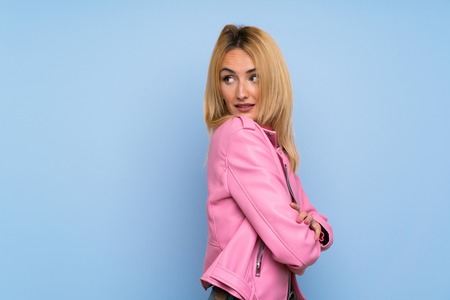 Young blonde woman with pink jacket over isolated blue background with arms crossed and happyの写真素材