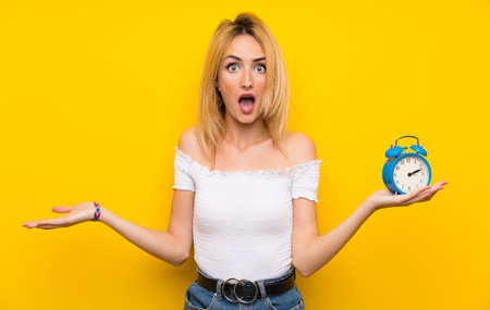 Young blonde woman over isolated yellow wall holding vintage alarm clock