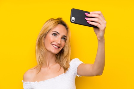 Young blonde woman over isolated yellow wall making selfie with cellphone