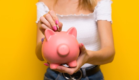 Young blonde woman over isolated yellow wall holding a big piggybankの写真素材