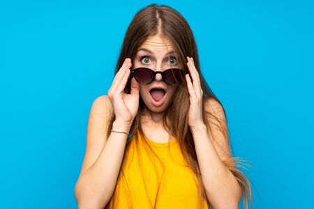 Young woman with long hair over isolated blue wall with glasses and surprised